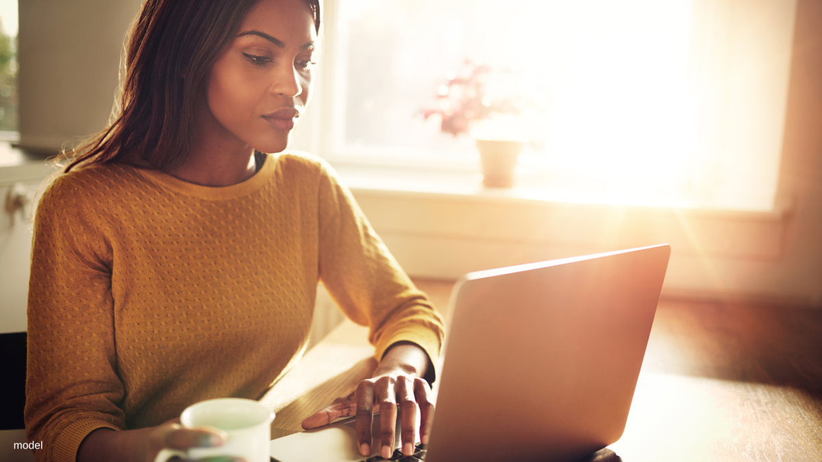 Model sitting at table with coffee and laptop.