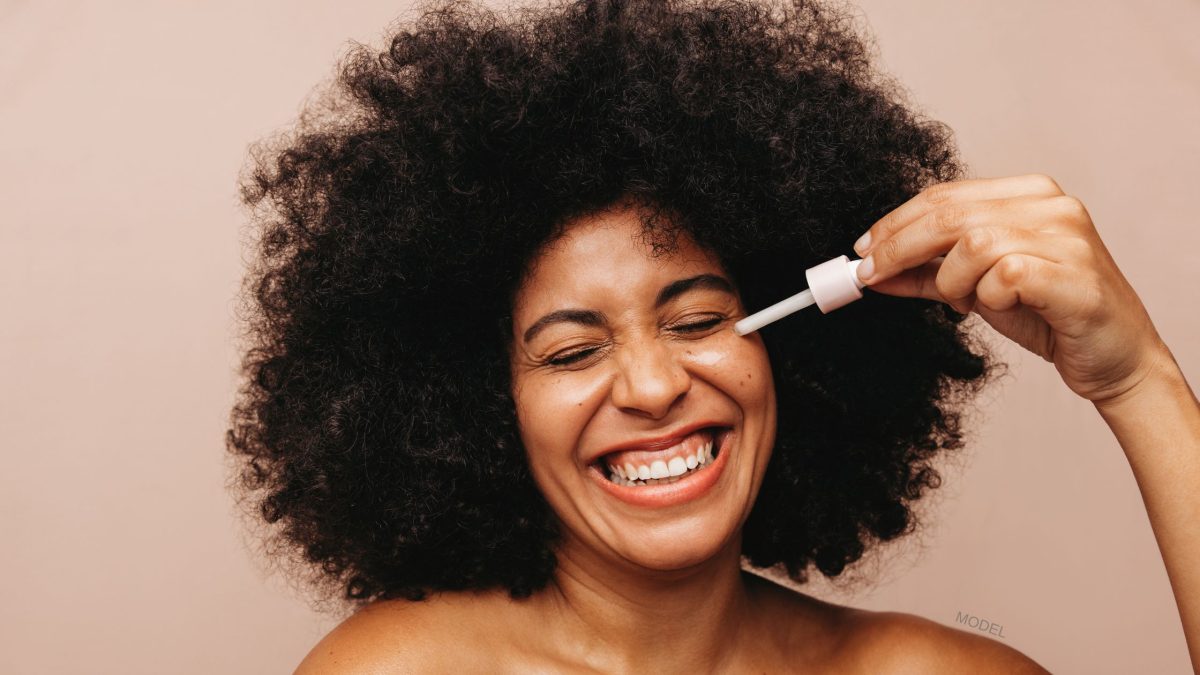 Woman smiling while she applies skincare to her face. (MODEL)