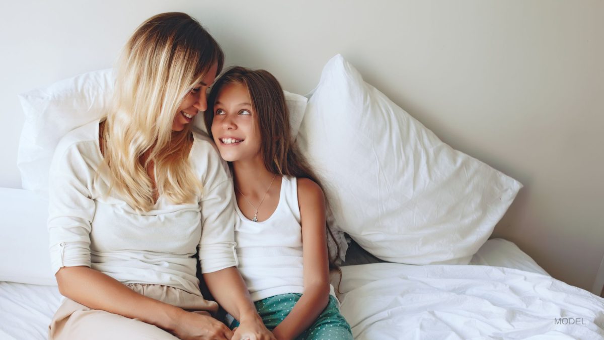 Mother and daughter sitting on bed and smiling at each other. (Models)