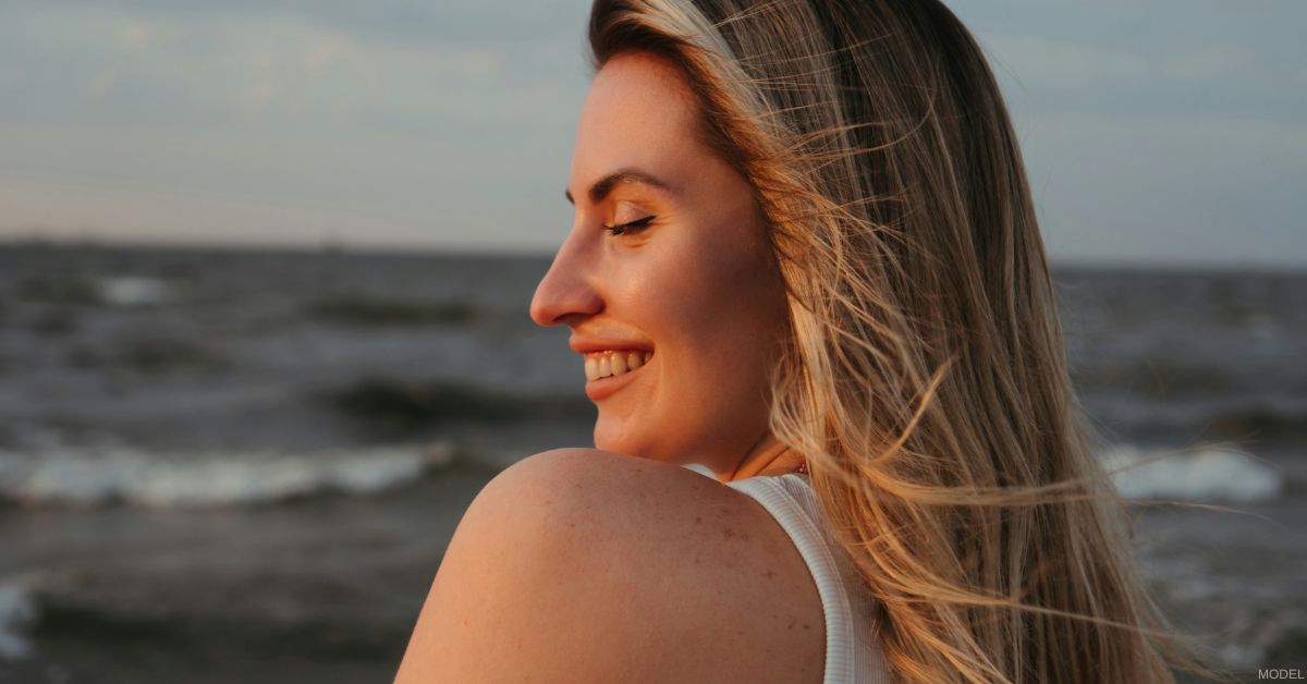 Woman smiling with eyes closed, standing by the ocean at sunset (model)
